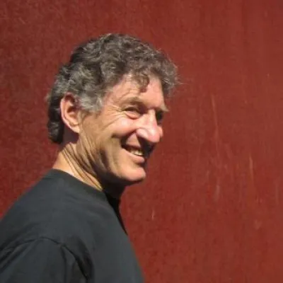 Steve Badanes stands in front of a red wall, facing to the right and smiling. He has short, curly gray hair and is wearing a black shirt.