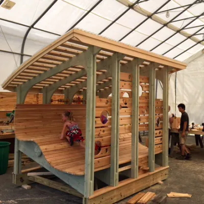 A curved wooden play structure is under construction inside a large tent workshop, with several adults working and a young child climbing on it.