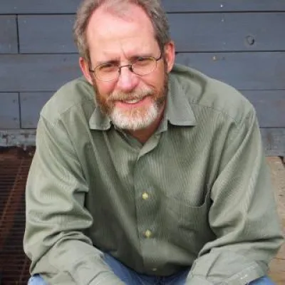 Sucosh Norton sits on an outside deck in front of a house. He is smiling at the camera, wearing glasses and a green button-down shirt.