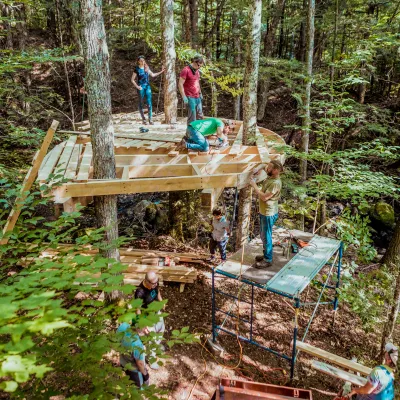 A group of seven people working on a wooden treehouse platform in the forest. Some people are standing on the ground, some are on a set of scaffolding and the rest of the group are standing on the wooden platform.