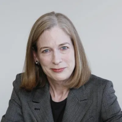 Sylvia Smith smiles at the camera against a grey background. She has shoulder length brown hair and is wearing a gray blazer.