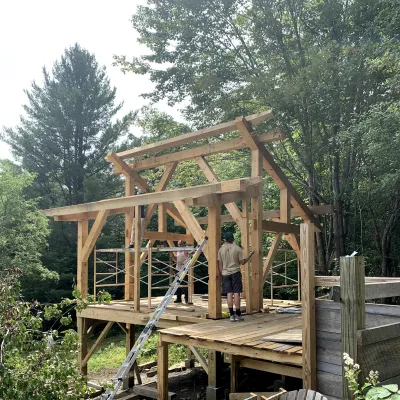 A small timber frame structure, with leafy trees and an adirondack chair in the foreground.