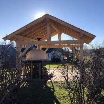 A timber frame over a pizza oven stands in a lawn. Blue sky and sun in the background.