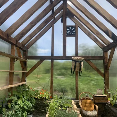 Interior shot of a timber frame greenhouse. Farm is visible outside of the clear walls and roof, and there are plants inside.