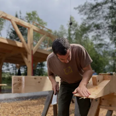 A student brushes sawdust off a fresh chisel cut, with a timber frame in the background.