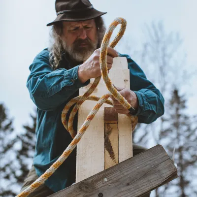 A man wearing a blue shirt and brimmed hat uses rope to lash timbers together.