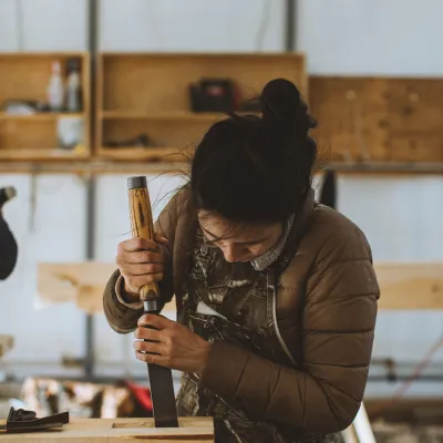 A student with dark hair in a bun and overalls uses a chisel on the end of a timber.