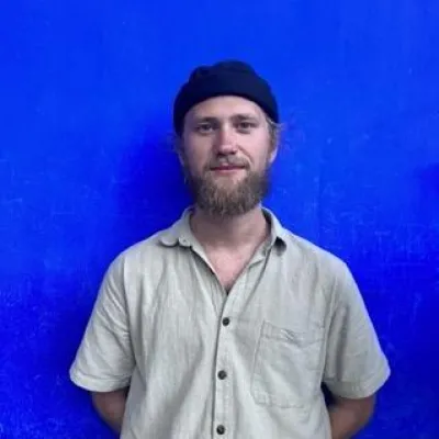 Tinder Kiely stands in front of a plain royal blue background. He is wearing a beige, short-sleeved button down shirt and black beanie.