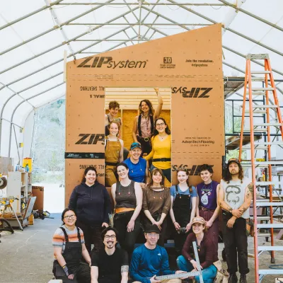 Group photo of the tiny house class in front of their framed and sheathed tiny house. They are smiling at the camera in an outdoor classroom.
