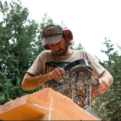 Tom uses a borer to make holes in a timber. He is wearing a grey t-shirt, grey baseball hat, and orange ear protection.
