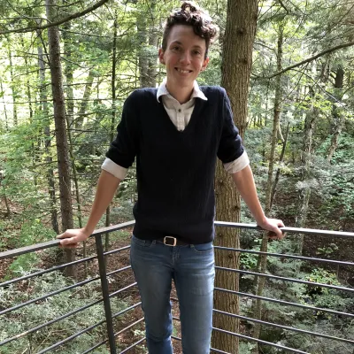 Trish stands in a treehouse against the railing. She has short, brown hair and is wearing jeans and a black sweater.