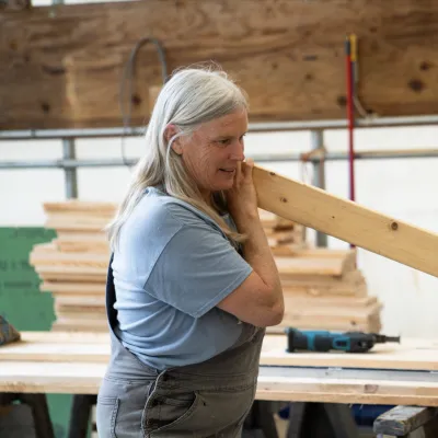 A woman wearing overalls sights down a two-by-four piece of lumber.