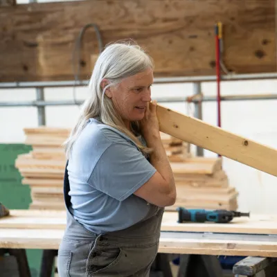 A person with long gray hair and overalls sights down a piece of lumber