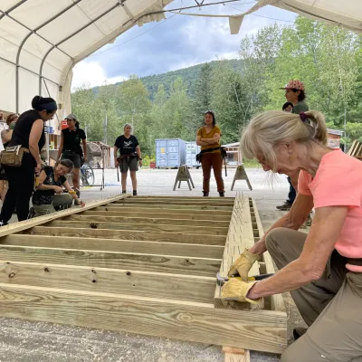 A group of students builds a floor deck. A woman in a pink shirt is hammering nails in the foreground.
