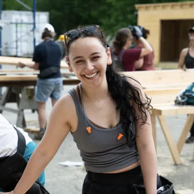 A woman smiles at the camera. She is wearing a grey tank top and has long dark hair in a ponytail. She is wearing orange earplugs around her neck and leaning over a piece of lumber.