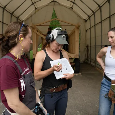 Students gather around an instructor holding plans in an outdoor classroom