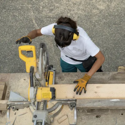 An aerial shot looking down at a student with brown hair and wearing a white t-shirt using a chop saw