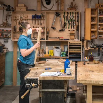 Two women at work benches facing each other measure pieces of wood in the woodshop