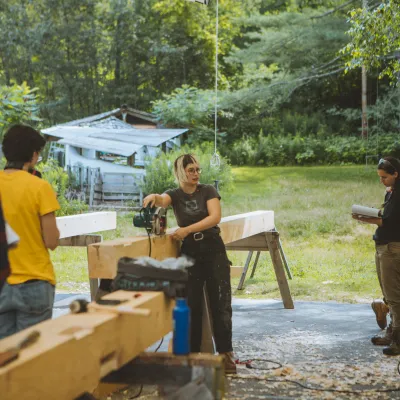 An instructor speaks to a group of students in an outdoor classroom, with cut timbers in the foreground.