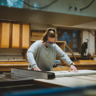 A person wearing a grey shirt and hair in a ponytail makes a cut on the table saw.
