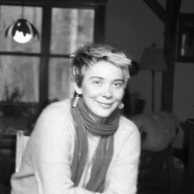 Wynne Hannan sits inside in front of a window. The black and white image shows Wynne with short hair, smiling, and wearing a scarf.