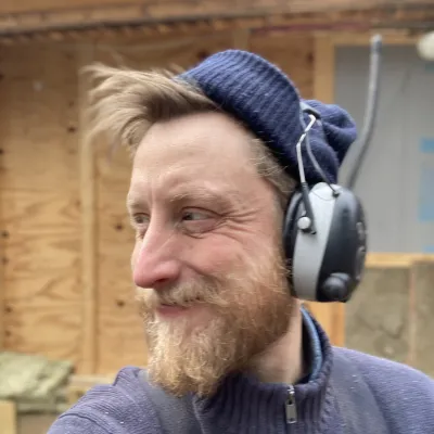 Zach is photographed outside on a jobsite. He is looking to the left, wearing ear protection and a blue beanie.