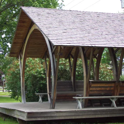 A pavilion in a public green space with a unique curved structure and shingled roof.
