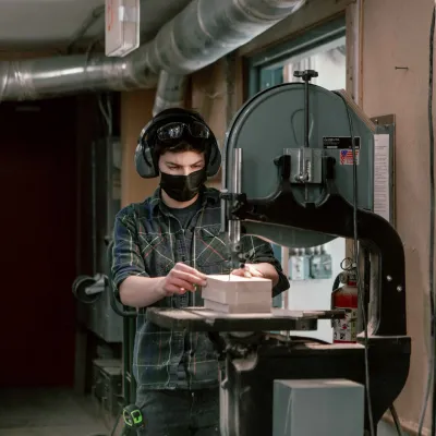 A student with short dark hair and gray flannel shirt uses the bandsaw in the woodshop.