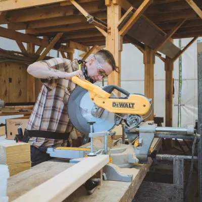 A person with buzzed hair uses a chop saw to cut lumber in a woodshop.