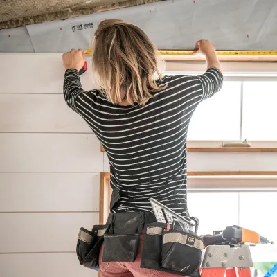 A person with blonde hair and black-and-white striped shirt uses a measuring tape on the top of a door frame