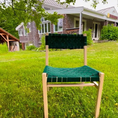 A maple chair with green woven seat and back is pictured outside in front of the main building on the lawn with tree branches overhead.