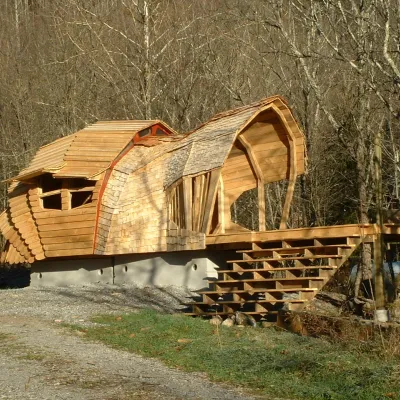A uniquely shaped wooden structure with shingle siding and a curved roof, built on a concrete foundation with stairs and a deck in a wooded area.