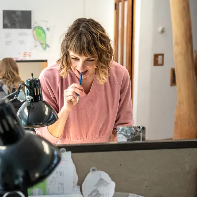 A person with medium blonde hair and wearing a pink shirt looks intently at work on a drafting table