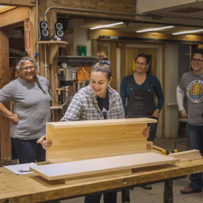 An instructor holds a piece of wood on a work bench, demonstrating for students standing behind.