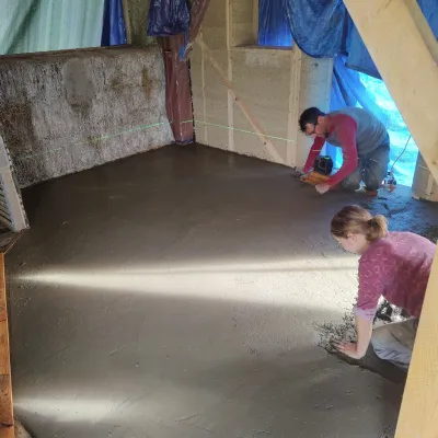 A construction site of a strawbale house covered in temporary tarps with two people kneeling and applying a dark gray earthen floor.