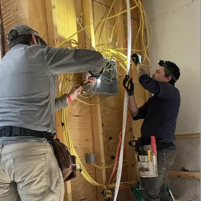 Two people work on an electrical panel in a framed building.