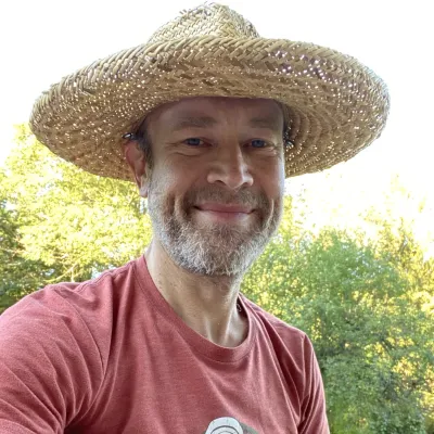 George smiles at the camera, photographed outside wearing a red shirt and wide-brimmed straw hat and holding a guitar 