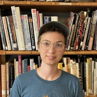 Grace smiles at the camera in front of shelves of books. They have short brown hair and brown eyes, and are wearing a baseball cap and blue shirt with a gold logo of a home that says "tiny house fest vermont"