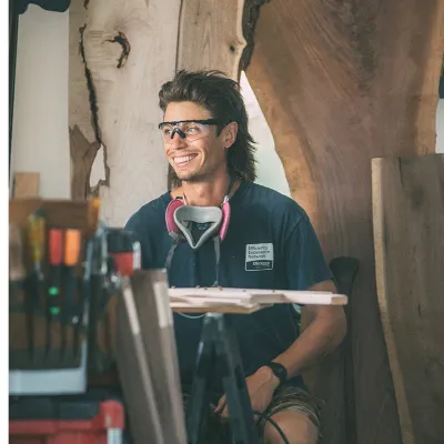 A smiling woodworker wearing safety glasses sits in a workshop surrounded by live-edge wood slabs and tools.