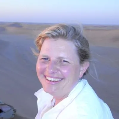 Janine smiles at the camera outside. There is blue sky behind her and her blonde hair is pulled back. She is wearing a white button-down shirt.