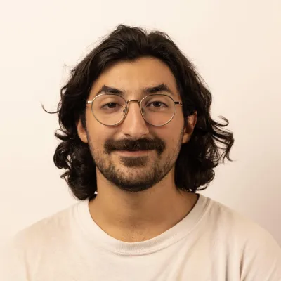 Leo smiles at the camera. He has dark hair, a beard and glasses. He's wearing a white t-shirt and background is white. 