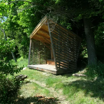 A wooden pavilion with slatted siding and built in bench on a wooded trail in the summer.