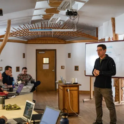 Shot of a class in a lecture in the main studio. Students are sitting facing the projector screen, and the teacher is standing in front of the screen speaking. 