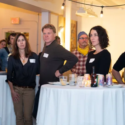People stand and listen to an off-camera speaker at an event. They are wearing nametags and standing around bar tables with white tablecloths.