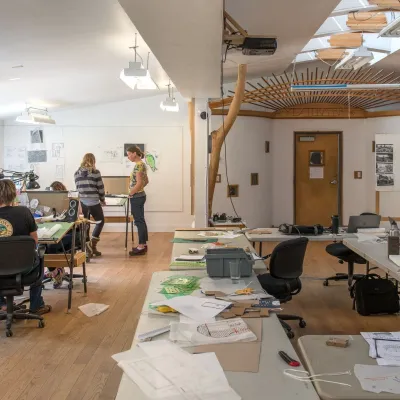 A wide shot of the yestermorrow main studio, with students busy at drafting tables and mingling around the room. The center table is in the foreground scattered with handouts.