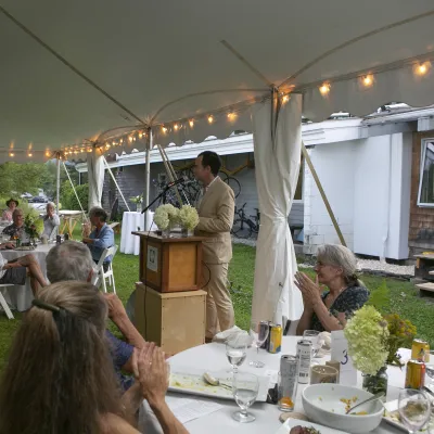 Britton stands at a podium addressing tables of guests under a white tent.