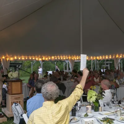 A man in the foreground raises a program at a seated event under a tent