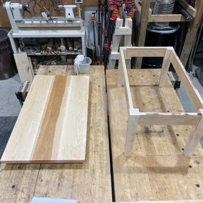A maple table top sits on a work bench next to assembled legs and rails.