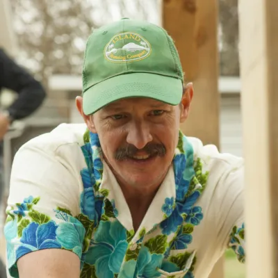 Man with a large mustache, wearing a Hawaiian shirt and baseball cap, looks forward. 