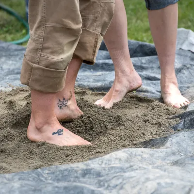 A tarp on the ground, covered in earthen materials with two sets of feet mixing the materials.